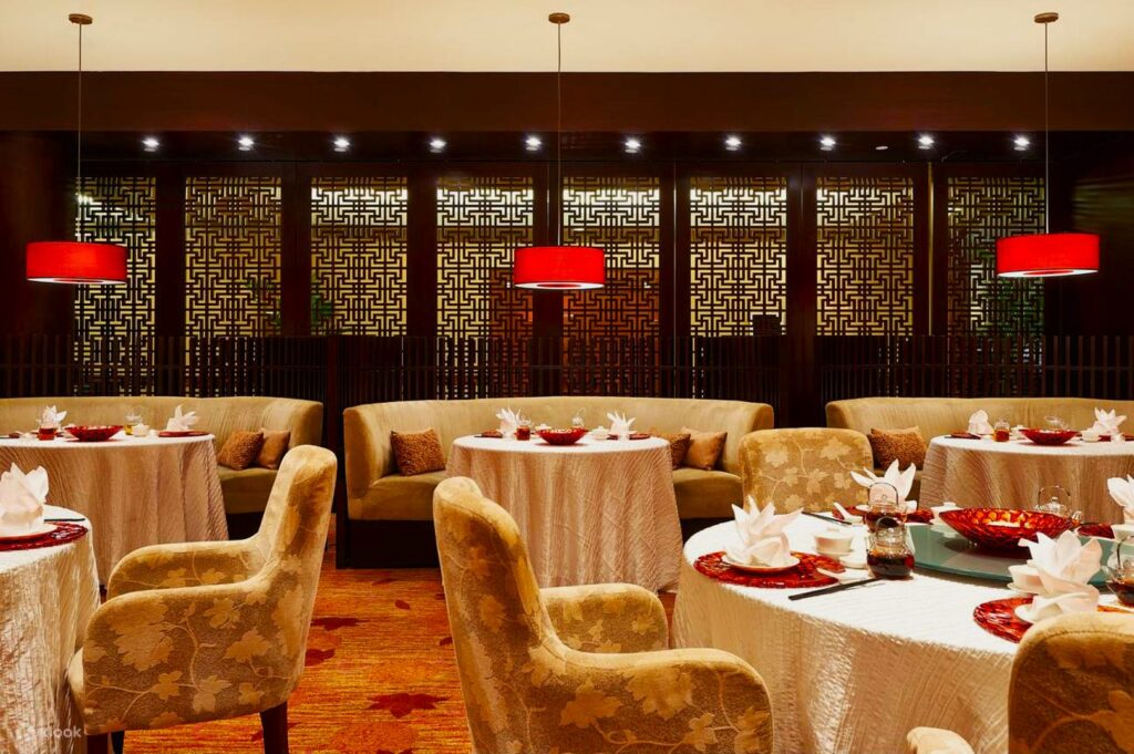 Elegant restaurant interior with round tables draped in white cloth, set with red plates and folded napkins. Plush beige chairs and red hanging lamps add warmth.
