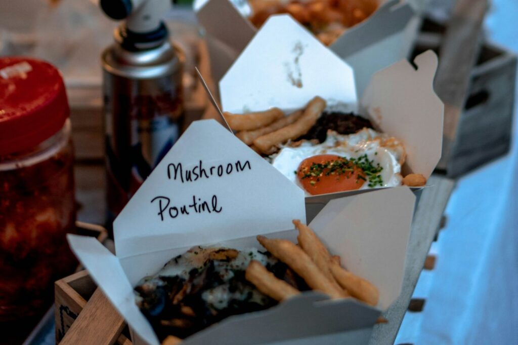 Open takeout boxes display Mushroom Poutine and fries, topped with fried eggs and herbs. A can and jar are blurred in the background. Casual vibe.