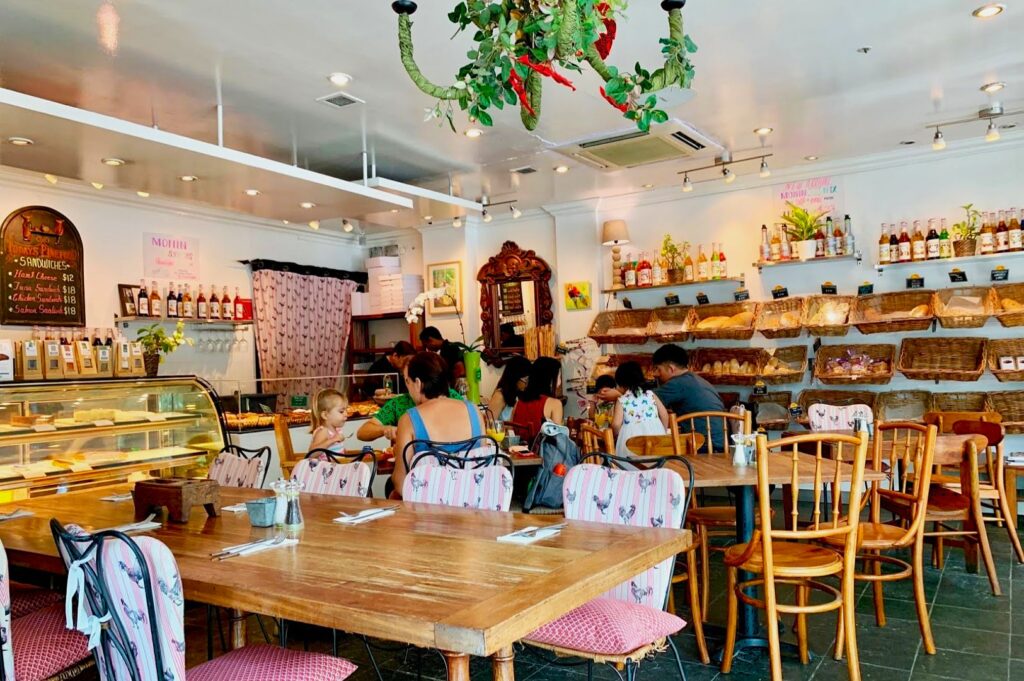 Cozy bakery interior with wooden tables, floral chairs, and a green chandelier. Shelves display bread and bottles, with patrons dining casually. Warm, inviting atmosphere.