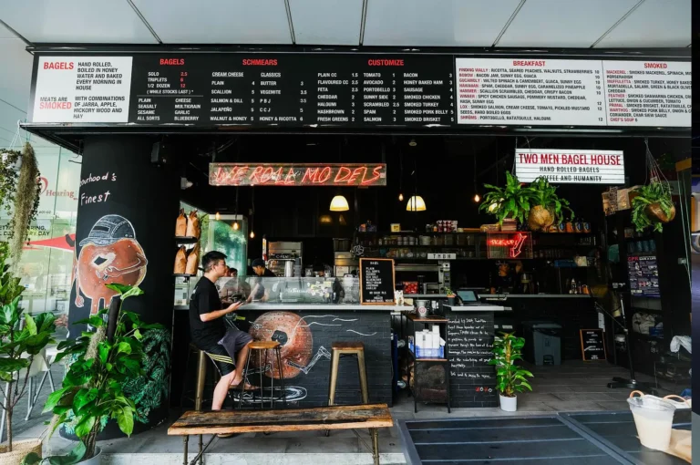 A cozy bagel shop with a blackboard menu displays various options. A person sits at the counter on a stool. Lush plants add a vibrant touch.