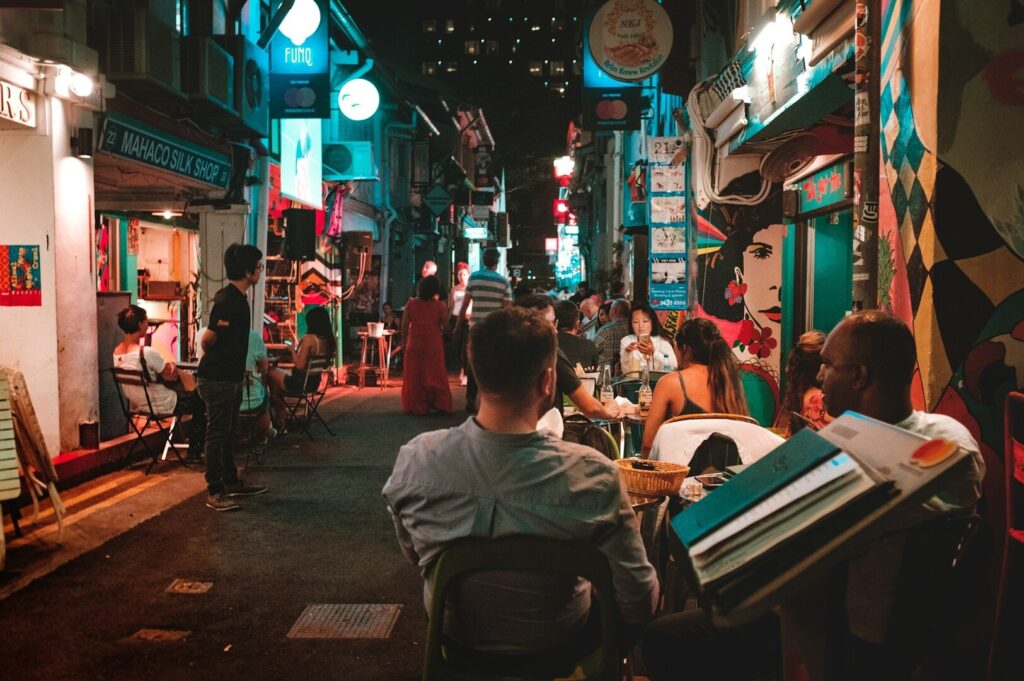 A lively, narrow street at night with people dining outdoors, surrounded by vibrant, colorful murals and neon signs, creating a bustling atmosphere.