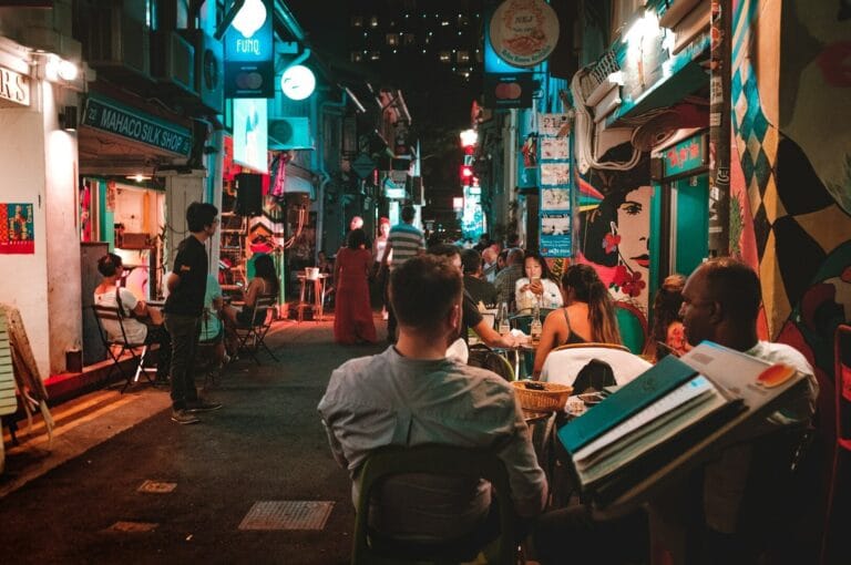 A lively, narrow street at night with people dining outdoors, surrounded by vibrant, colorful murals and neon signs, creating a bustling atmosphere.
