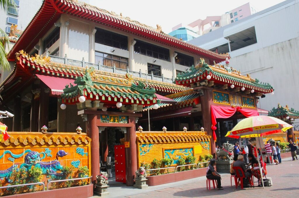 A vibrant Chinese temple with ornate red and green roofs, detailed dragon motifs, and gold accents. People sit under a red and yellow umbrella nearby.