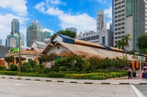 A traditional market building with a sloped roof is surrounded by lush greenery in an urban setting, with skyscrapers and a clear blue sky in the background. People walk along the sidewalk, adding a lively atmosphere.