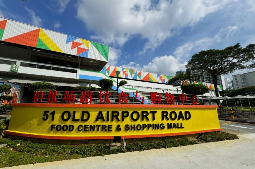 Bright, colorful facade of 51 Old Airport Road Food Centre & Shopping Mall. Clear blue sky, lush trees, and geometric patterns create a vibrant atmosphere.