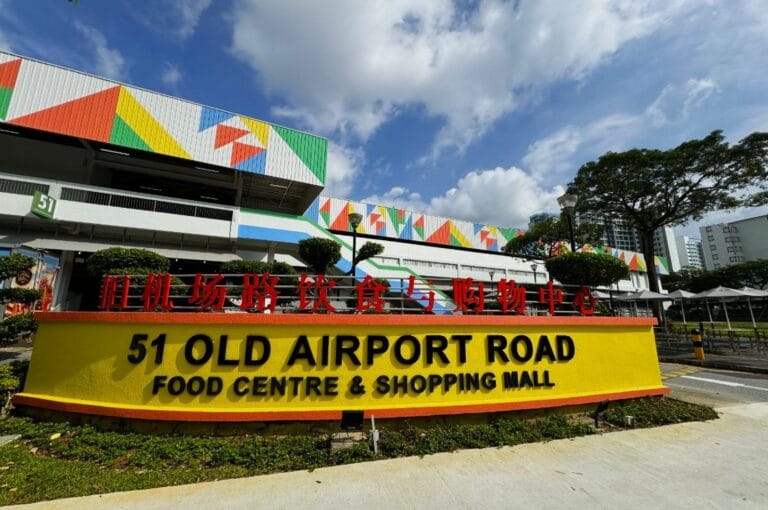 Bright, colorful facade of 51 Old Airport Road Food Centre & Shopping Mall. Clear blue sky, lush trees, and geometric patterns create a vibrant atmosphere.