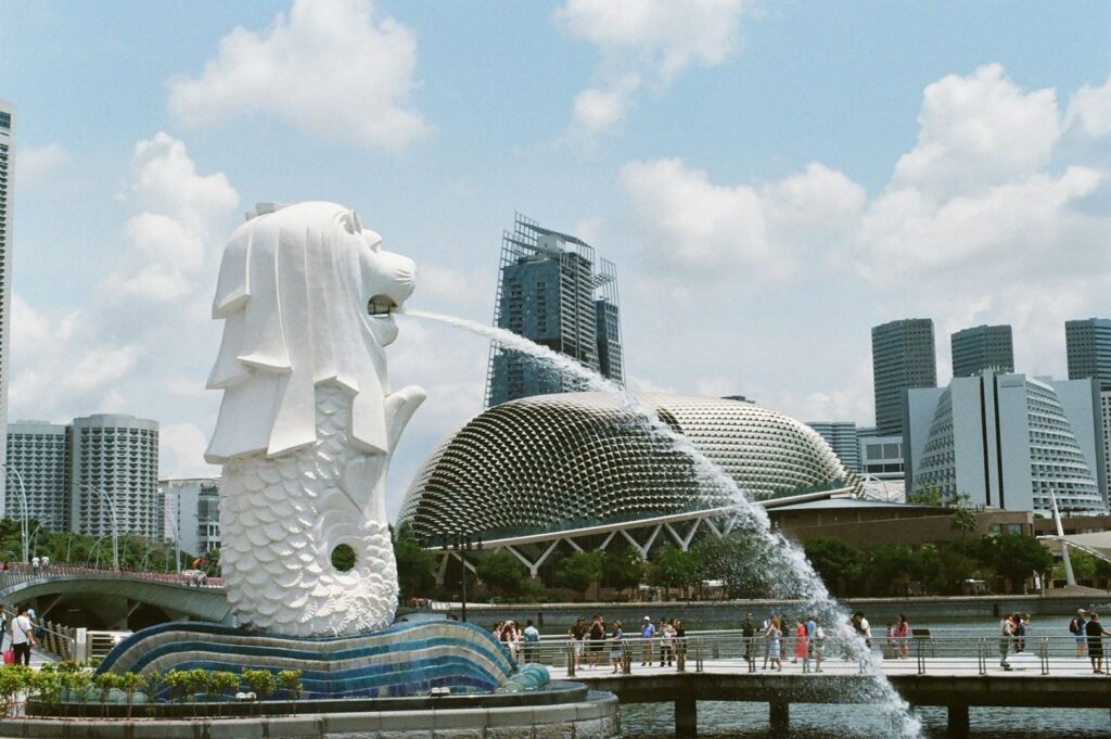 Merlion statue in Singapore spouts water against a backdrop of modern skyscrapers and the domed Esplanade. Tourists gather on a sunny day.