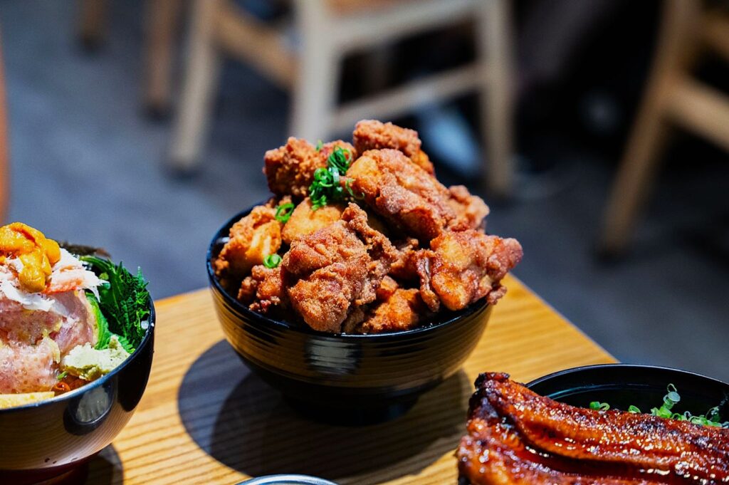 A black bowl filled with golden-brown fried chicken topped with green herbs sits on a wooden table, surrounded by other vibrant dishes in a cozy setting.