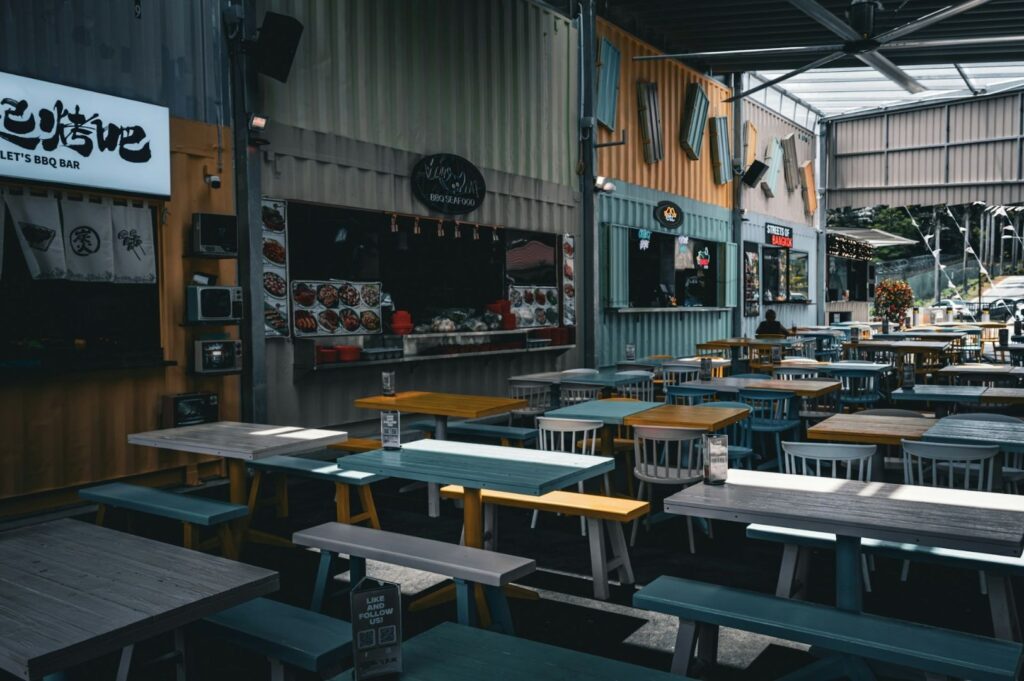 Outdoor dining area with colorful picnic tables and chairs, surrounded by food stalls in repurposed shipping containers. The setting is casual and inviting.