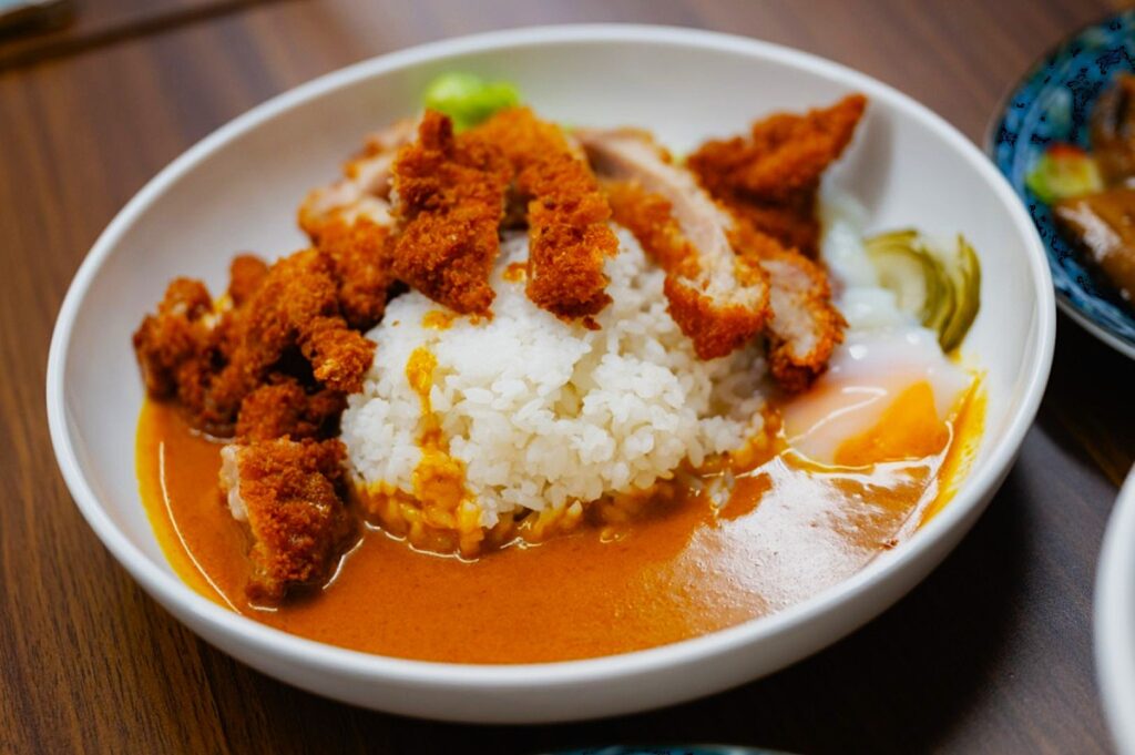 A white bowl with Japanese curry rice topped with golden fried chicken cutlets. A soft-boiled egg and vegetables are on the side. Warm and inviting.