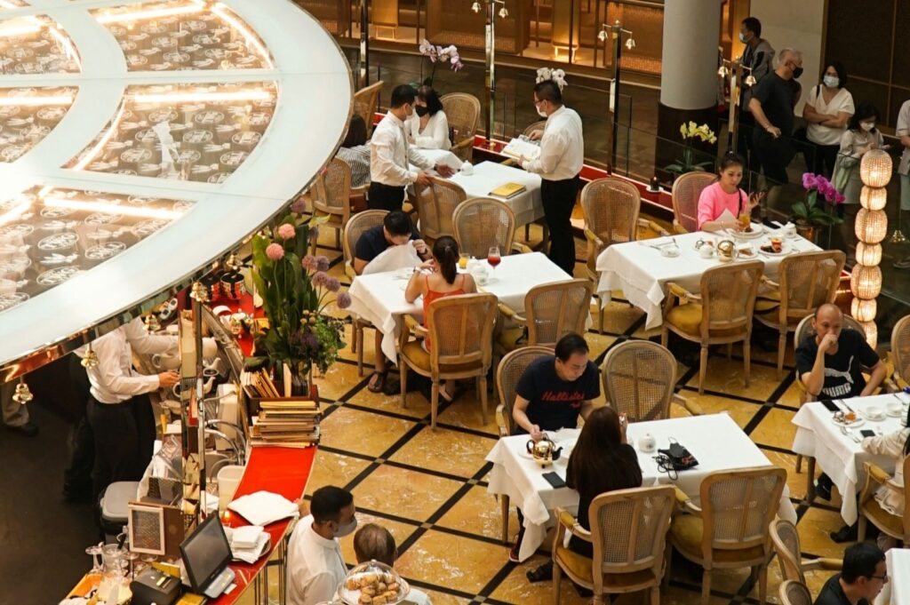 Overhead view of an elegant restaurant filled with patrons at white-clothed tables. Waitstaff in uniforms actively attend to guests, creating a lively, sophisticated atmosphere.