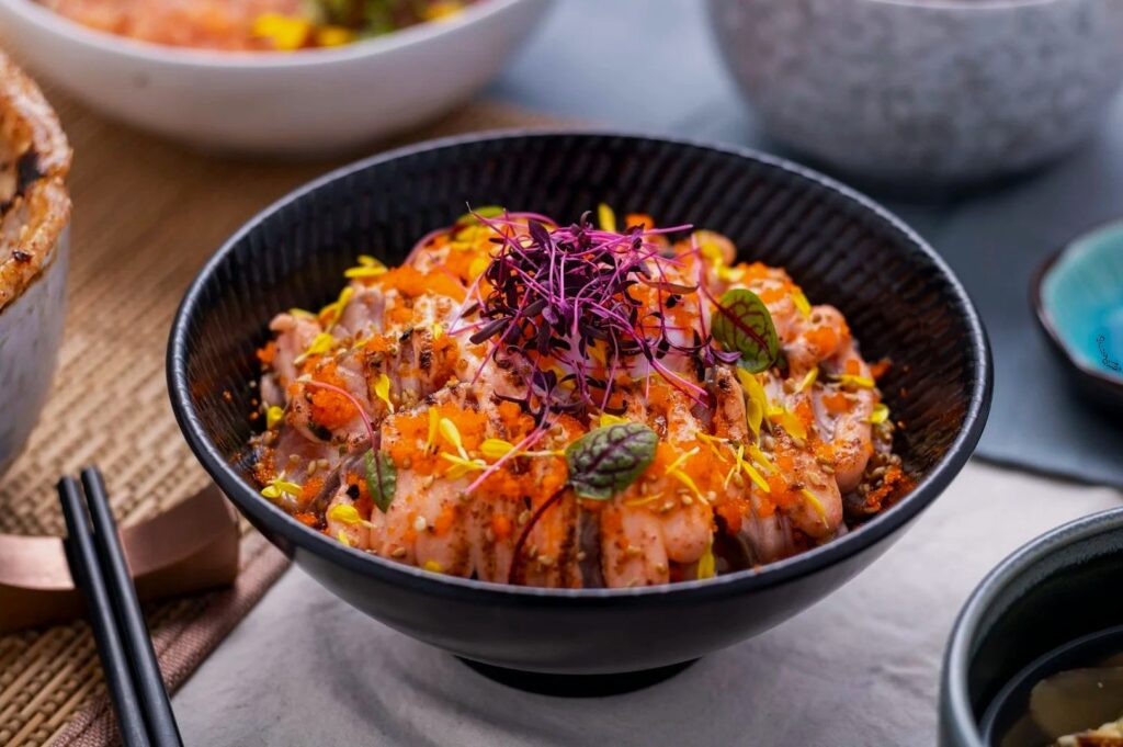 A black bowl filled with seasoned, seared salmon, topped with vibrant microgreens and orange sauce. It sits on a table with chopsticks, evoking a fresh, appetizing feel.