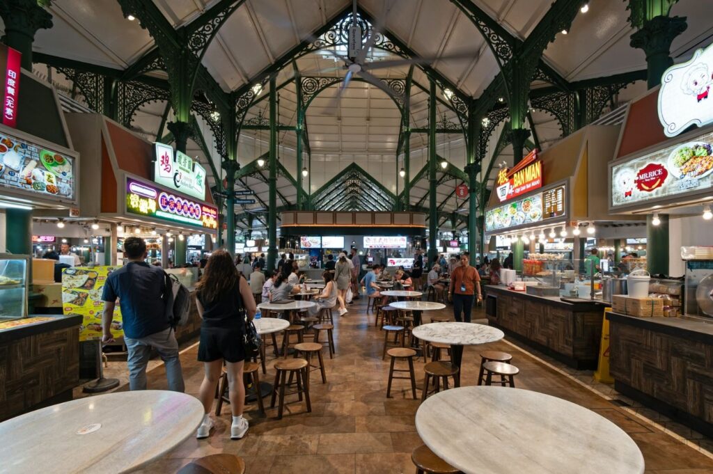 Bustling food hall with ornate green architecture; people walk and dine at round tables amidst colorful food stalls, creating a lively atmosphere.