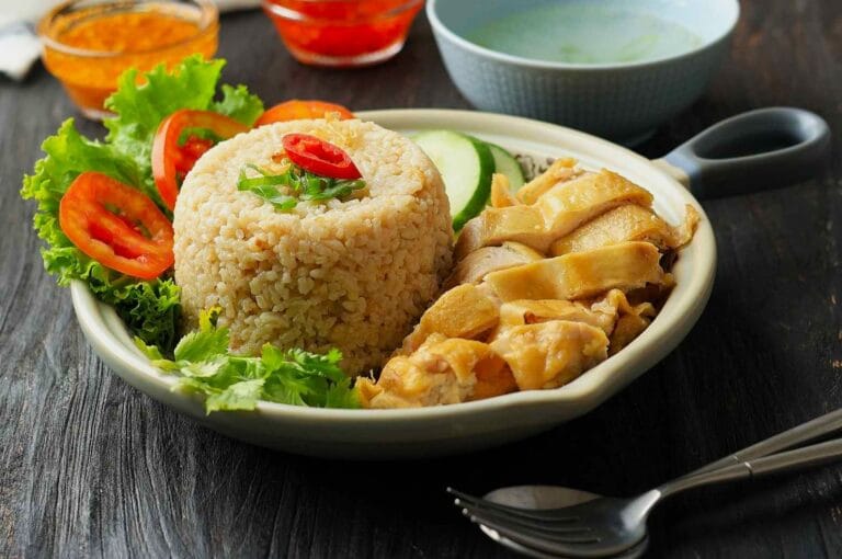 A bowl of Hainanese chicken rice with sliced chicken, rice, lettuce, cucumber, and tomato. Sauces in bowls and a spoon are visible. Inviting and fresh.