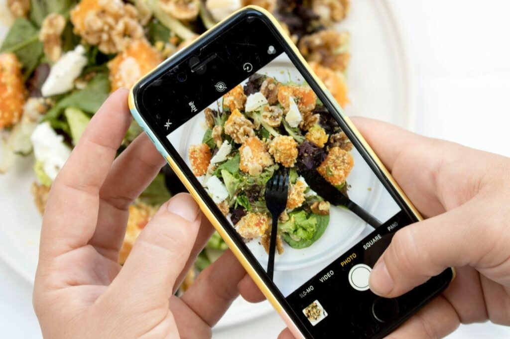 A person captures a photo of a colorful salad with chicken, greens, and cheese on a smartphone, emphasizing food photography and modern dining experiences.