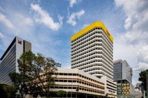 Cityscape with modern buildings featuring angular designs and yellow accents under a clear blue sky. Trees and clouds add depth, creating a vibrant urban scene.