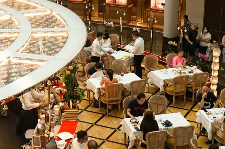 Aerial view of a busy, elegant restaurant. Diners seated at white-clothed tables, attended by staff in white uniforms. Warm lighting and floral decor.
