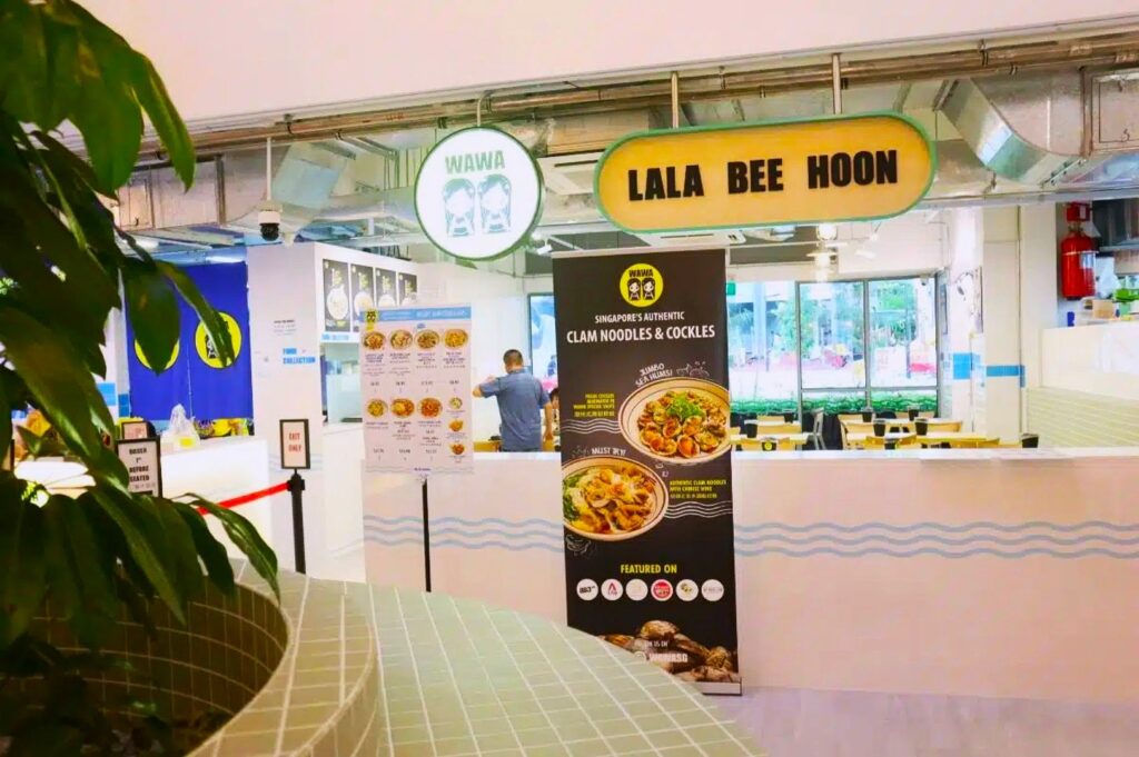 Interior of an eatery with a sign reading "Lala Bee Hoon." A poster features clam noodles, and a few diners can be seen in the bright, inviting space.