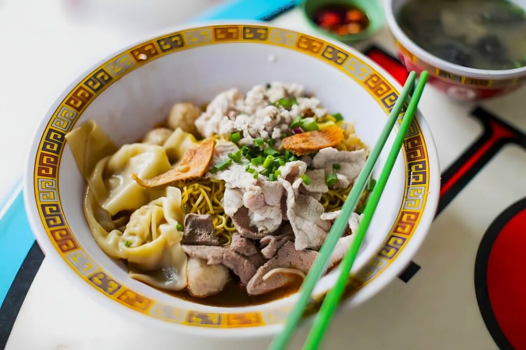A bowl of noodle soup with sliced pork, dumplings, meatballs, and green onions. Green chopsticks rest on the bowl, evoking a sense of warmth.