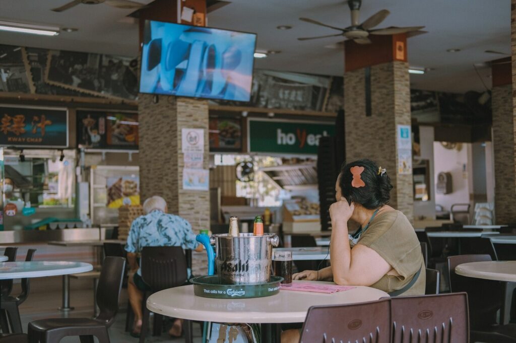 A person with a butterfly hair clip sits alone in a food court, looking at a ceiling-mounted TV. The scene is quiet, with empty chairs around.