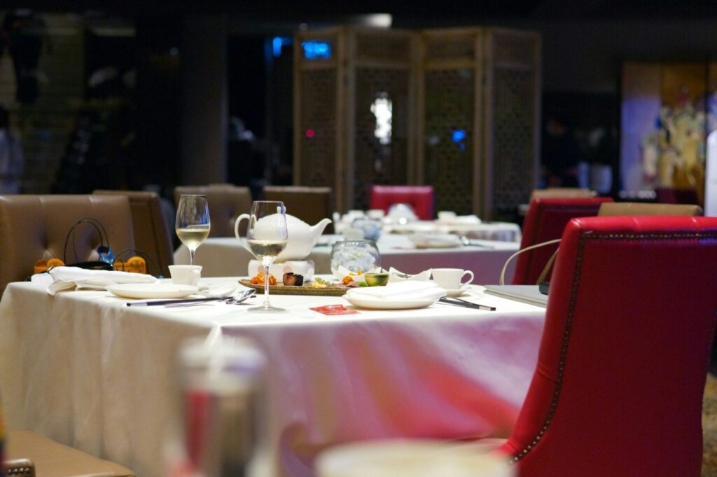 Elegant restaurant interior with tables set for dining. White tablecloths, red chairs, wine glasses, and plates create a refined, intimate atmosphere.