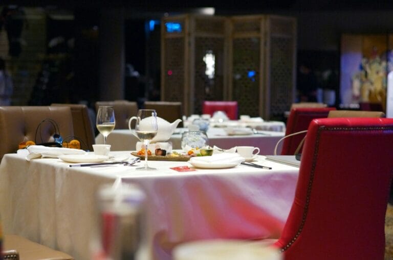 Elegant restaurant interior with tables set for dining. White tablecloths, red chairs, wine glasses, and plates create a refined, intimate atmosphere.