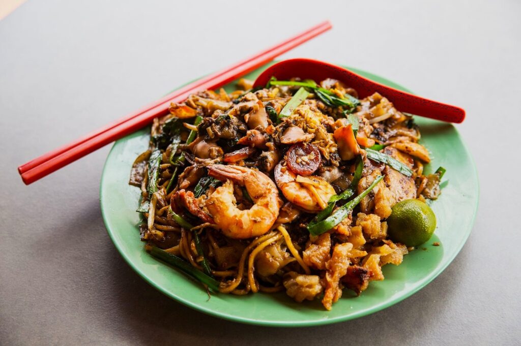 A vibrant plate of stir-fried noodles topped with shrimp, vegetables, and greens on a green plate. A red spoon and chopsticks rest beside it, exuding a savory and appetizing aroma.