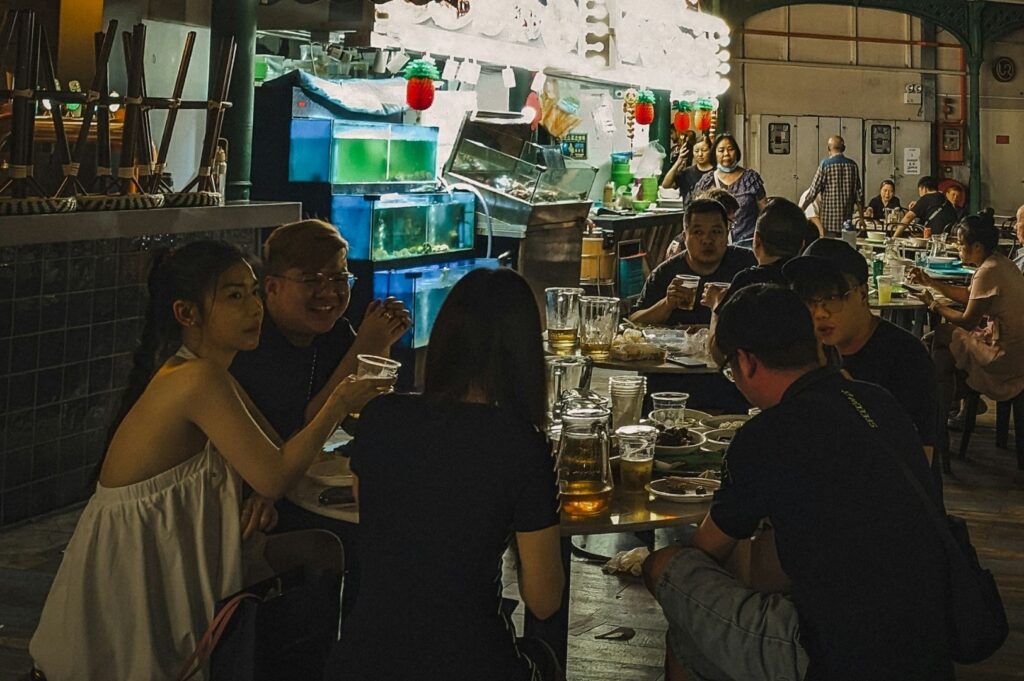 A group of people sit around a table in a bustling, dimly lit restaurant. They are enjoying drinks and food, evoking a warm, lively atmosphere.