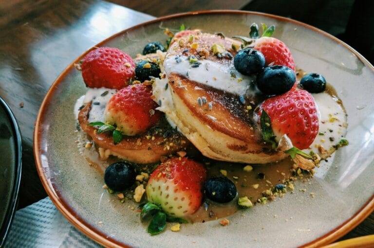 A plate of fluffy pancakes topped with fresh strawberries, blueberries, and crushed pistachios, drizzled with creamy sauce, creating a colorful and inviting breakfast scene.