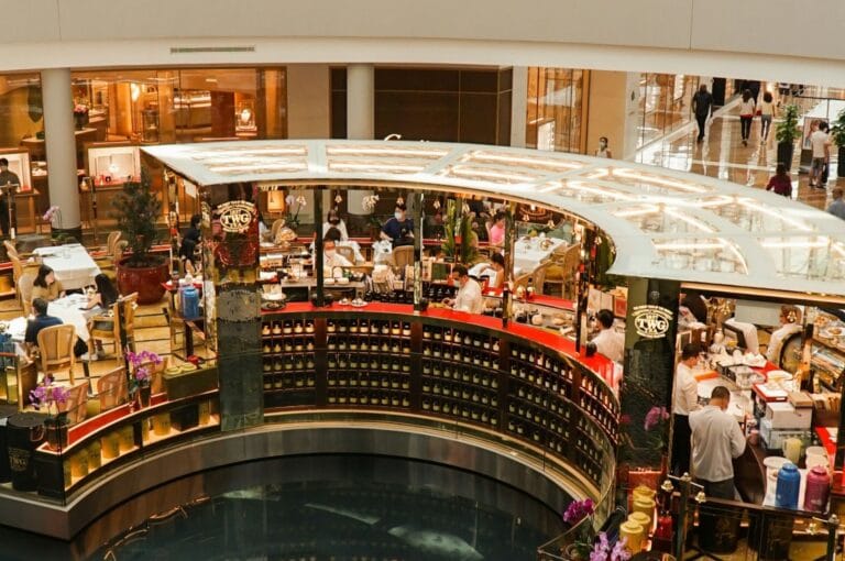 Elegant shopping mall interior with a circular cafe surrounded by bustling shoppers. The cafe features a bright counter and decorative plants, creating a lively atmosphere.