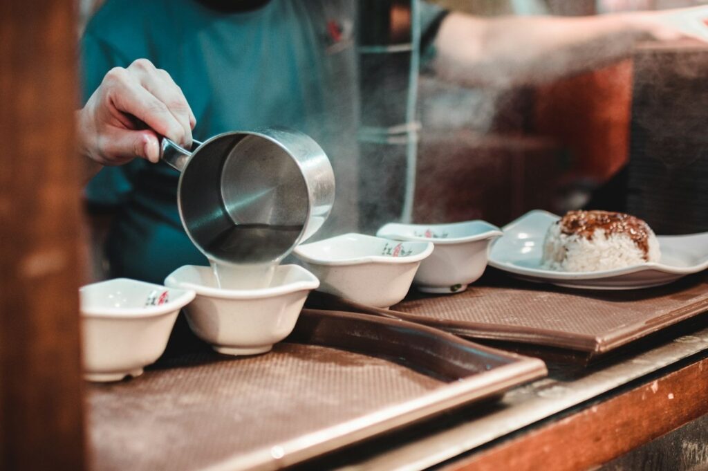 A person pours hot liquid into white bowls on a tray. Steam rises, with a plate of rice nearby. The scene conveys warmth and culinary preparation.