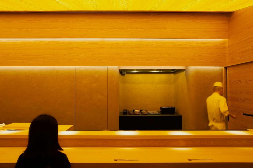 A minimalist sushi restaurant interior with warm wooden tones. A person sits at a counter while a chef in white attire prepares food, creating a serene ambiance.