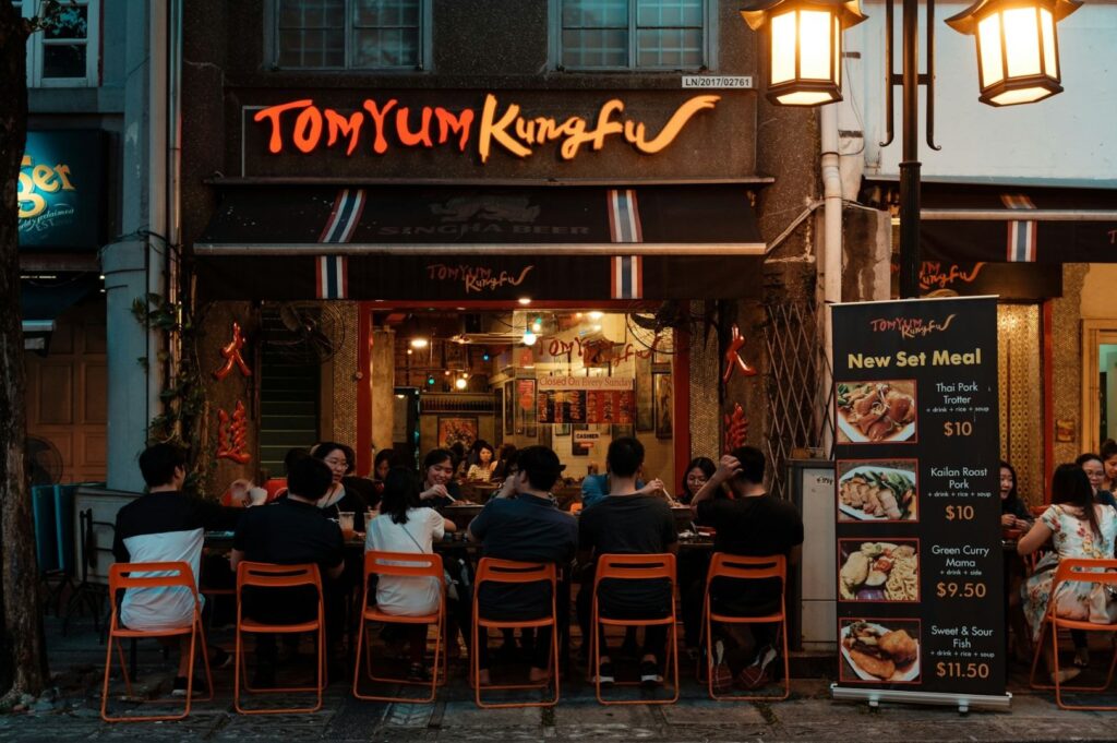 Outdoor evening dining scene at a Thai restaurant, "Tom Yum Kung Fu." Guests sit on orange chairs at a communal table. A menu board promotes set meals.