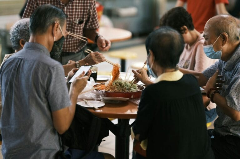 A group of elderly people sits around a table, sharing a meal with chopsticks. The atmosphere is warm and social. One person wears a face mask.