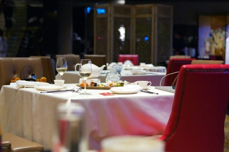 Elegant restaurant scene with a white tablecloth and red chairs. The table is set with wine glasses, a teapot, and neatly arranged plates, creating a cozy ambiance.