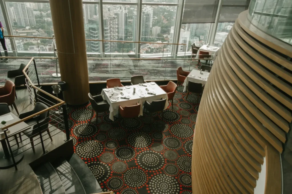 A modern restaurant with large windows overlooking a cityscape. Elegant tables are set on a patterned carpet, creating a sophisticated ambiance.