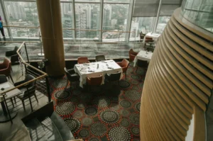 A modern restaurant with large windows overlooking a cityscape. Elegant tables are set on a patterned carpet, creating a sophisticated ambiance.