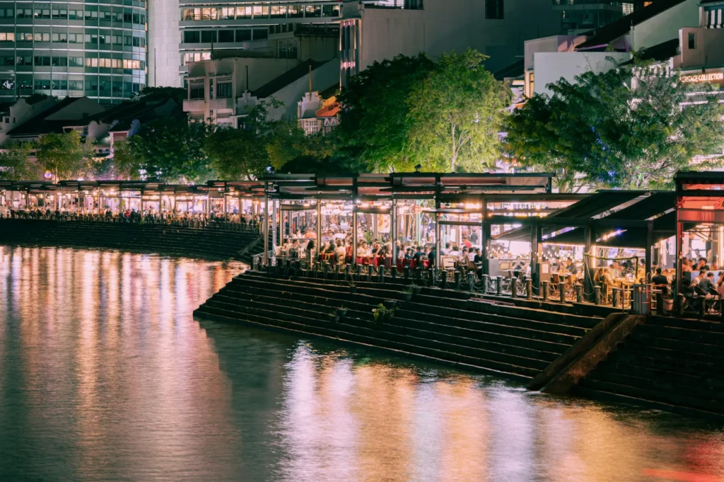 A vibrant riverside scene at night with bustling restaurants, bright lights, and reflections on the water. Trees and modern buildings in the background.