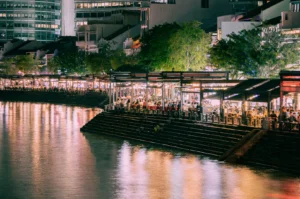 A vibrant riverside scene at night with bustling restaurants, bright lights, and reflections on the water. Trees and modern buildings in the background.