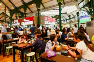 Bustling food court with diverse people dining at long tables under high, ornate ceilings. Bright signs and food stalls create a lively atmosphere.