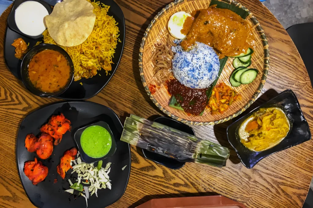 An array of colorful dishes on a wooden table, featuring biryani, spicy chicken, nasi lemak with blue rice, curry, and vibrant accompaniments.