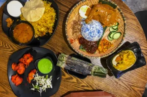 An array of colorful dishes on a wooden table, featuring biryani, spicy chicken, nasi lemak with blue rice, curry, and vibrant accompaniments.