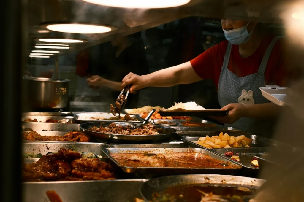 Vendor in a red shirt and apron serves food from trays under warm lights. The scene is bustling, filled with colorful, appetizing dishes.