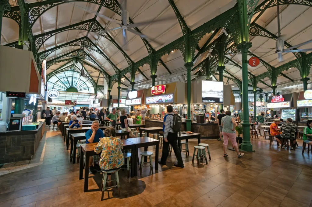 A bustling food court with ornate green iron columns and high ceilings. People sit at wooden tables, enjoying diverse eateries and a lively atmosphere.
