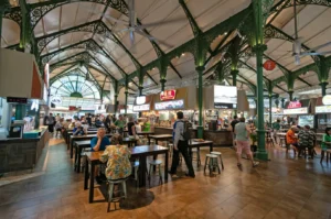 A bustling food court with ornate green iron columns and high ceilings. People sit at wooden tables, enjoying diverse eateries and a lively atmosphere.