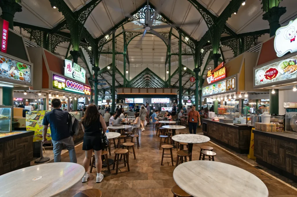 A bustling indoor food court with ornate green metal beams, various food stalls lining the sides, and people walking or sitting at round tables.