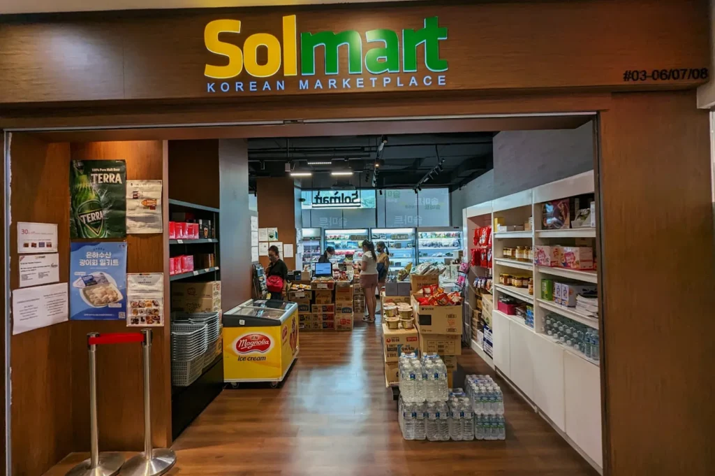 Entrance to Solmart, a Korean marketplace, with shelves of groceries, bottled water, and a checkout counter in view. Bright, inviting atmosphere.