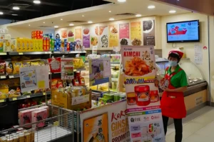 A grocery store with Korean food items like kimchi on promotion. Shelves display snacks and groceries. A staff member in red uniform stands beside a sale poster.