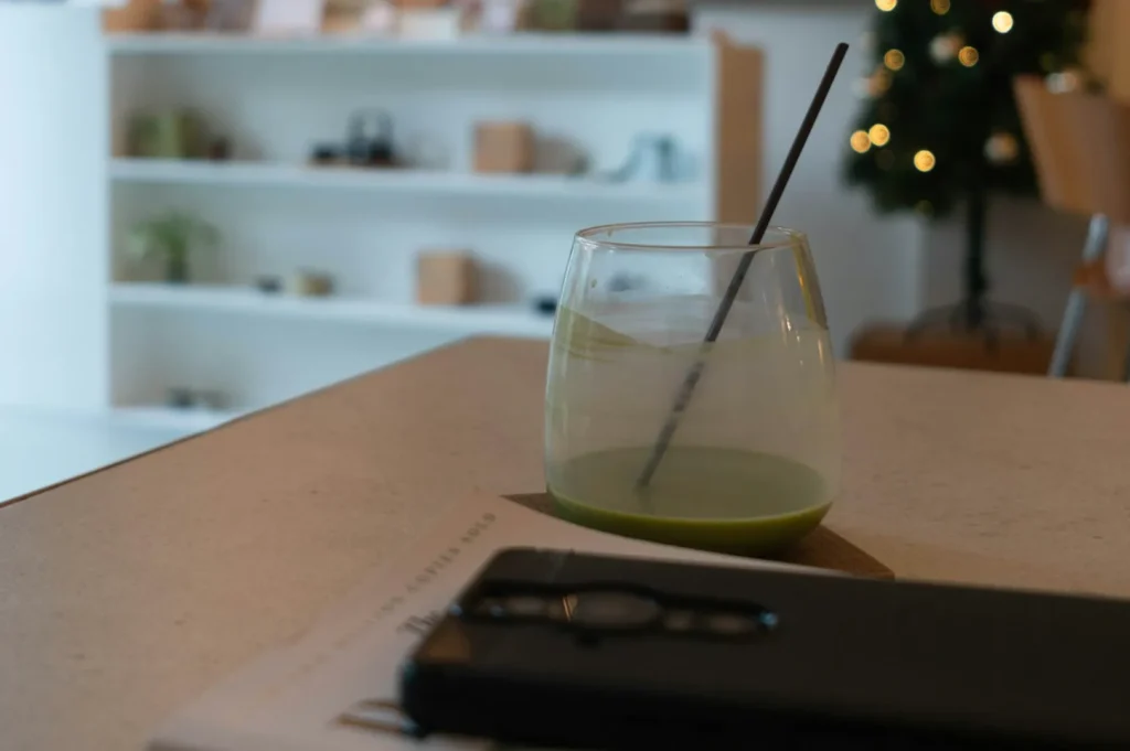 A glass of green juice with a straw sits on a table next to a smartphone and papers. The background features blurred shelves and a lit Christmas tree.
