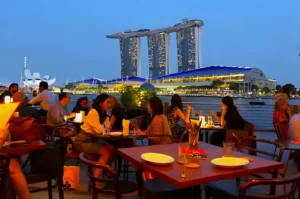 People dine on a rooftop terrace at dusk, enjoying a view of the illuminated Marina Bay Sands in Singapore. The ambiance is lively and relaxed.